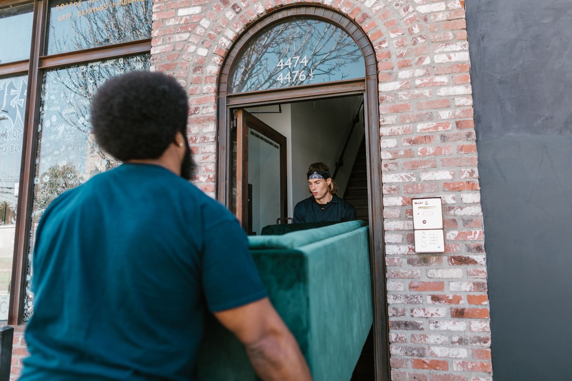 Two adults carrying a sofa through the doorway of a brick house