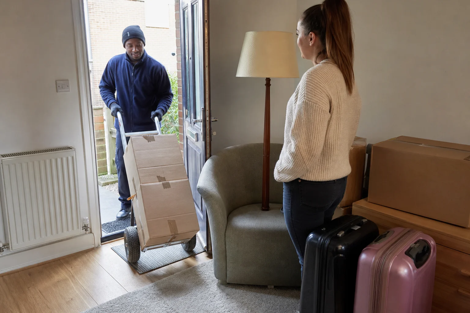 A removal worker wheeling packed boxes from a compact UK flat while the customer waits with an armchair and suitcases